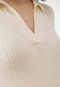 Top tricot beige clair avec un col en V, présentant une texture côtelée verticale et un col doux. Le tissu semble lisse et élastique.