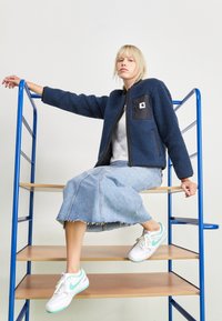 Navy fleece jacket with black pocket, paired with a denim skirt and white sneakers featuring mint accents. Model seated on blue ladder.