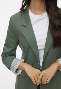 Blazer verde realizzato in tessuto morbido, con un design su misura, rever a punta e maniche arrotolate con un motivo a righe.