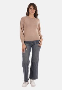 Maglione beige lavorato a maglia con maniche lunghe, scollatura rotonda e una consistenza morbida, abbinato a jeans a gamba larga scuri e sneakers bianche.