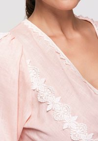 Femme portant un chemisier rose pâle avec une bordure florale en dentelle blanche le long d'un décolleté en V et un détail bouffant aux épaules.