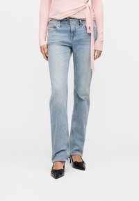 Γυναίκα που φοράει ανοιχτό μπλε τζιν κανονικής γραμμής, μαύρες μυτερές γόβες με φιόγκους και μακρυμάνικο ροζ τοπ τύπου wrap σε λευκό φόντο.