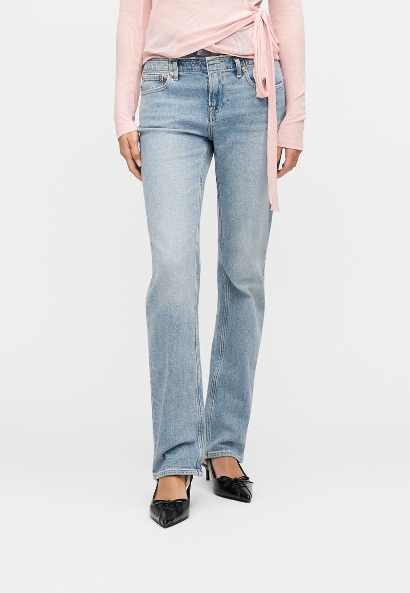 Γυναίκα που φοράει ανοιχτό μπλε τζιν κανονικής γραμμής, μαύρες μυτερές γόβες με φιόγκους και μακρυμάνικο ροζ τοπ τύπου wrap σε λευκό φόντο.