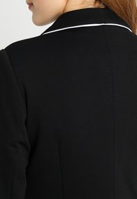 Czarna marynarka z klasycznym kołnierzem z białym wykończeniem, o strukturalnym kroju i gładkiej fakturze materiału. Widoczne detale szwów.