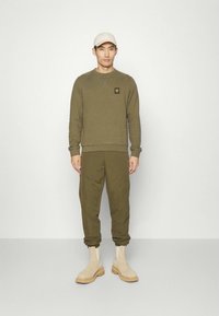 Sweatshirt verde oliva com um patch de logótipo, combinado com calças de treino olive e botas ankle bege, exibido contra um fundo simples.