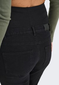 Tumši zaļas augsta vidukļa džinsi ar gludu jostasvietu, divām aizmugurējām kabatām, daļēji redzama teksturēta olīvu zaļa augšdaļa.