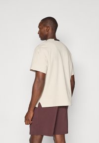 T-shirt beige à manches courtes avec une coupe décontractée et des fentes sur les côtés, associé à un short bordeaux foncé. Le tissu semble doux et léger.