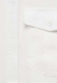 Gros plan sur une poche de chemise blanche avec un rabat fermé par un petit bouton blanc sur un fond de tissu uni.