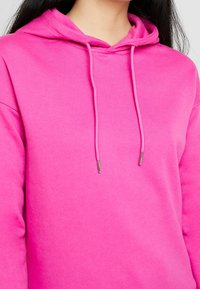 Růžová mikina vyrobená z měkké tkaniny, se stahovací kapucí a kovovými konci šňůrky, a s uvolněným střihem.