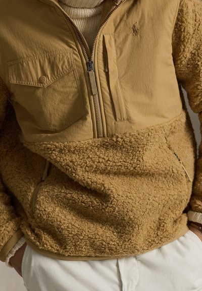 Μπεζ φλίς μπλούζα με μισό φερμουάρ, με υφή στο κάτω μέρος και λεία επιφάνεια στο πάνω, δύο τσέπες και λογότυπο στο στήθος.