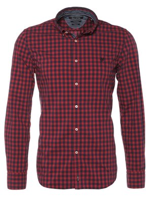 Homme portant une chemise à carreaux rouge et noir, un jean noir retroussé aux chevilles, des chaussures noires, une veste bleu marine sur l'épaule.