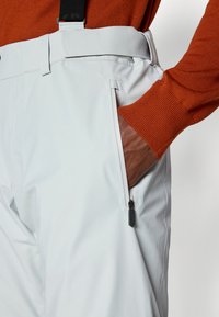 Voděodolné bílé kalhoty s kapsou na zip, spárované s hnědou rukou a oranžovým svetr. Mají nastavitelné šle.