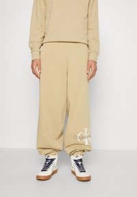 Beige katoenen sweatpants met een elastische taille en boorden, met een Calvin Klein-logo in lichtbruin bij de enkel. Gecombineerd met hoge sneakers.