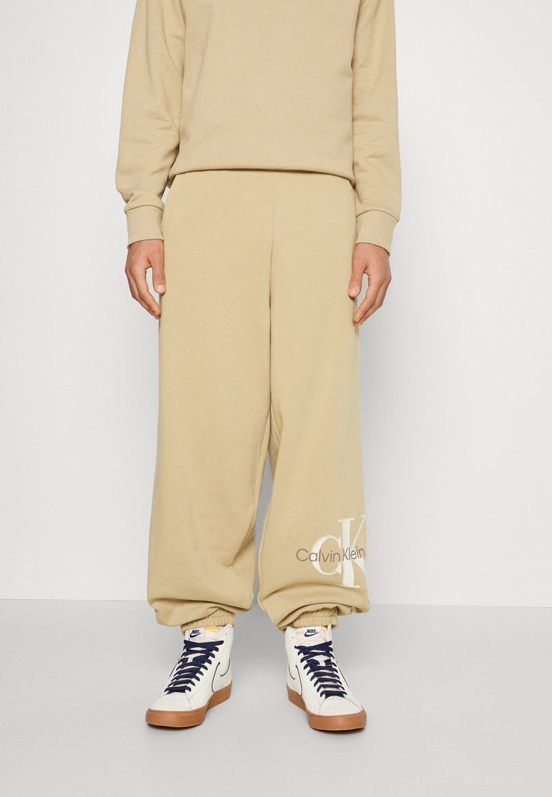 Beige katoenen sweatpants met een elastische taille en boorden, met een Calvin Klein-logo in lichtbruin bij de enkel. Gecombineerd met hoge sneakers.