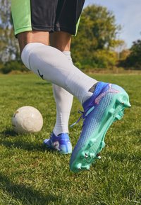 Blue and green soccer cleat with a textured surface, purple accents, and visible studs, worn with white socks and black shorts on grass.