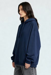 Donna con capelli lunghi e scuri, indossa una felpa oversize blu navy e pantaloni grigi, rivolta verso sinistra su uno sfondo chiaro e semplice.