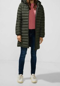 Polstrad grön lång kappa med huva, med horisontella kviltade linjer, kombinerad med en rosa topp, mörka jeans och vita sneakers.