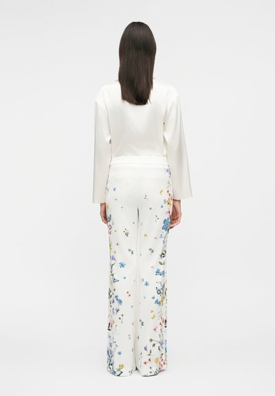 Γυναίκα με μακριά σκούρα μαλλιά που φοράει λευκό μακρυμάνικο τοπ και λευκό παντελόνι με φαρδιά μπατζάκια με πολύχρωμα λουλουδάτα σχέδια, που στέκεται με την πλάτη προς τα εμάς.