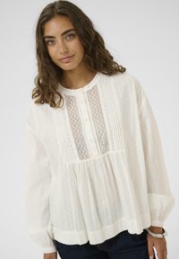 Femeie cu păr ondulat brunet, îmbrăcată cu o bluză albă cu mâneci lungi, detalii din dantelă și nasturi, asortată cu pantaloni închiși la culoare și o brățară argintie.