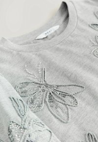 Tissu gris clair avec des motifs subtils d'abeilles en perles et sequins, doté d'un col rond et d'une étiquette de vêtement visible indiquant "NEXT".