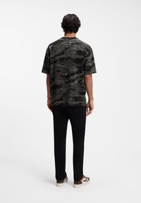 Άνδρας που στέκεται γυρισμένος με την πλάτη, φοράει μαύρο και γκρι πουκάμισο με μοτίβο και κοντά μανίκια, μαύρο παντελόνι και λευκά αθλητικά παπούτσια με πορτοκαλί λεπτομέρειες.