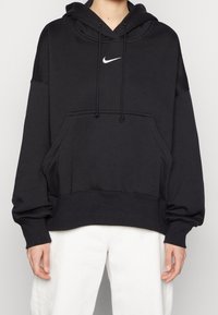 Haut à capuche noir avec une poche avant, logo Nike blanc, capuche avec cordon de serrage et poignets côtelés. Tissu doux, coupe lâche et design légèrement oversize.