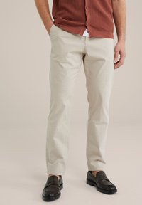Pantalones beige claro de corte recto y textura suave, combinados con mocasines oscuros. El conjunto incluye una camisa de botones de color óxido con textura acanalada.