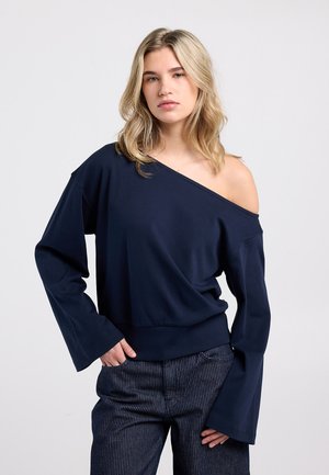 Marineblauwe off-shoulder top met lange mouwen en een ontspannen pasvorm, gemaakt van gladde stof; gecombineerd met broek met krijtstrepen.