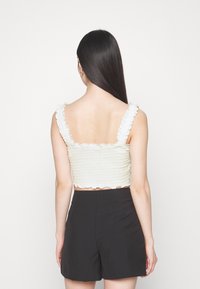 Cropped top in textured light green fabric with ruffled straps, featuring a wavy hem. Paired with tailored black shorts.