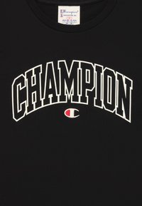 Must t-särk mustast puuvillast, millel on julge valge "CHAMPION" tekst kaarjas kujunduses, ja allosas väike punase ja valge värviga tikitud logo.