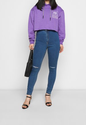 Sweat à capuche court violet avec imprimé graphique et cordons, associé à un jean skinny bleu taille haute avec des déchirures aux genoux et des talons noirs à lanières.