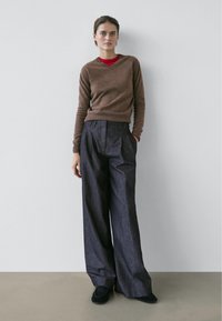 Pull marron à manches longues avec col en V, porté sur une chemise rouge, associé à un pantalon en denim foncé et ample. Porté avec des chaussures plates noires.