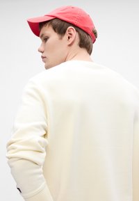 Jasnożółta bluza z luźnym krojem i prążkowanymi mankietami, w połączeniu z koralową czapką bejsbolową. Miękka tekstura, minimalistyczny design.