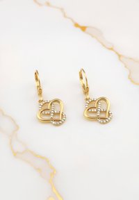 Orecchini a forma di cuore in oro con design intrecciato, accentuati da piccole pietre di rhinestone trasparenti. Finitura elegante su una superficie liscia.