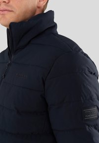 Marineblauer Puffermantel mit hohem Kragen, gestepptem Muster und einem linken Ärmelschild mit dem "Fundango"-Logo. Mit Reißverschluss vorne.