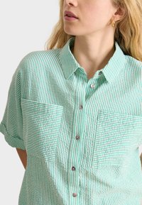 Short-sleeve, collared shirt in light green with horizontal white stripes. Features two textured front pockets and button closure.