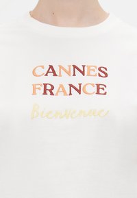 Hvit bomull t-skjorte med brodert tekst: "CANNES FRANCE" i oransje og brunt, og "Bienvenue" i gul kursiv.