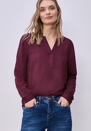 Ung kvinde med blondt hår iført en bordeauxrød bluse med lange ærmer og blå jeans, hænderne i lommerne, neutral baggrund.