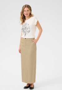 T-shirt blanc avec un design graphique noir, associé à une longue jupe beige avec un bouton sur le devant et des poches latérales. Ballerines noires.