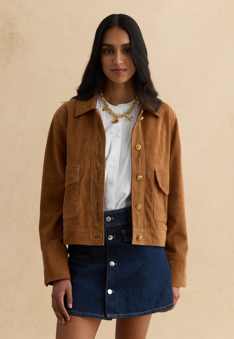 Mujer con chaqueta de ante marrón con botones dorados, blusa blanca, collar dorado y falda vaquera azul oscuro con botones, sobre fondo beige.
