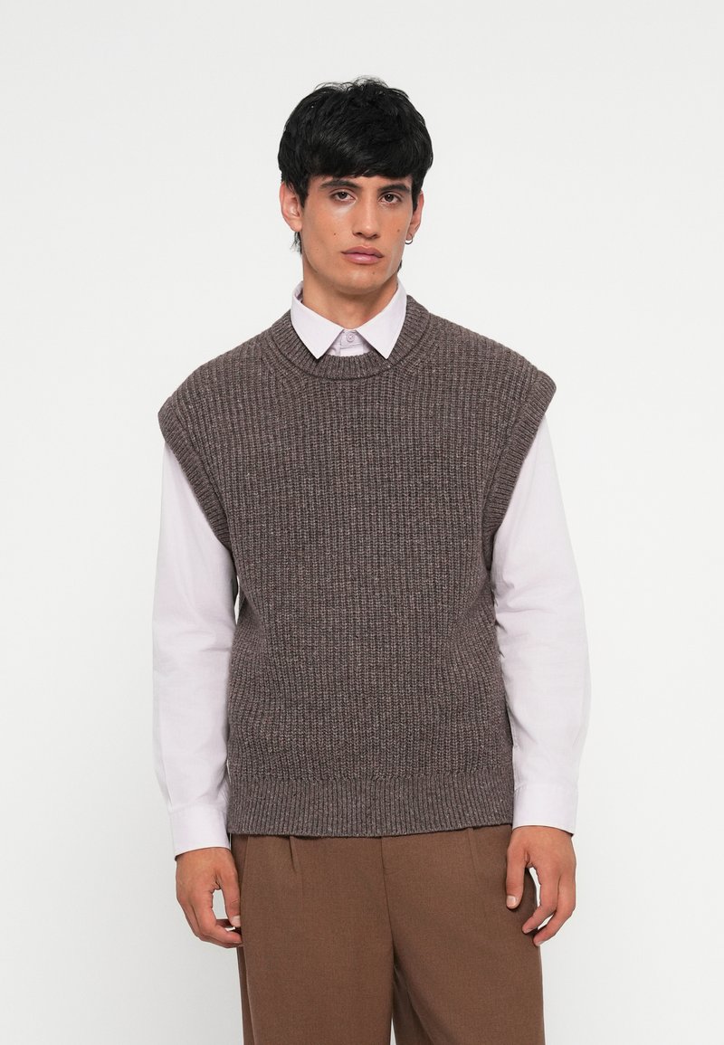 Gilet en maille marron avec une texture côtelée et un col rond. Porté par-dessus une chemise à col claire. Coupe décontractée, design sans manches.