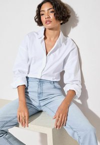 Camicia bianca con maniche arrotolate, abbinata a jeans azzurri a vita alta. Anelli dorati mettono in risalto le mani. Sfondo neutro.