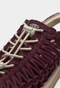 Κοντινό πλάνο σε βυσσινί πλεκτό σανδάλι με μπεζ κορδόνια από ελαστικό κορδόνι και πλαστικό κουμπί σε ανοιχτή σόλα.