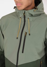 Veste étanche verte avec une fermeture éclair à l'avant, section inférieure en vert foncé contrasté et cordons de serrage jaunes sur la capuche. Comprend une poche sur la poitrine.