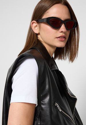 Donna con capelli castani lisci indossa occhiali da sole neri e un gilet di pelle nero sopra una camicia bianca a maniche corte su sfondo grigio.