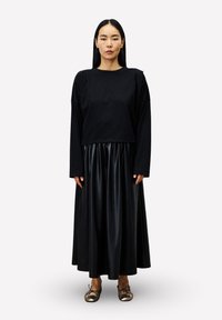 Zwarte top met lange mouwen en verlaagde schouders, gecombineerd met een plissé zwarte leren rok tot op de vloer en metalen instapschoenen.