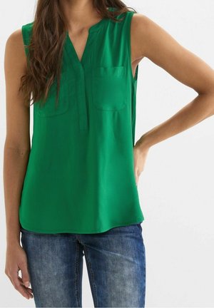 Kobieta ubrana w zieloną bluzkę bez rękawów z dwoma kieszeniami na piersi i niebieskie dżinsy, stoi z jedną ręką na biodrze na jednolitym tle.