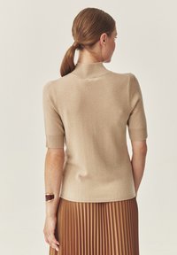 Maglietta in maglia beige a maniche corte con colletto alto, caratterizzata da una texture liscia. Il design posteriore presenta un delicato dettaglio di cucitura e una silhouette aderente.