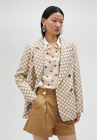 Dubbelknäppt jacka i beige med rutmönster, kombinerad med en krämfärgad skjorta med bruna prickar och högmidjade shorts i tan.