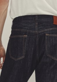 Tumman siniset farkut oransseilla ompeleilla, joissa on kaksi takataskua ja nahkapaikka vyötäröllä. Sileä pinta ja muotoiltu istuvuus.